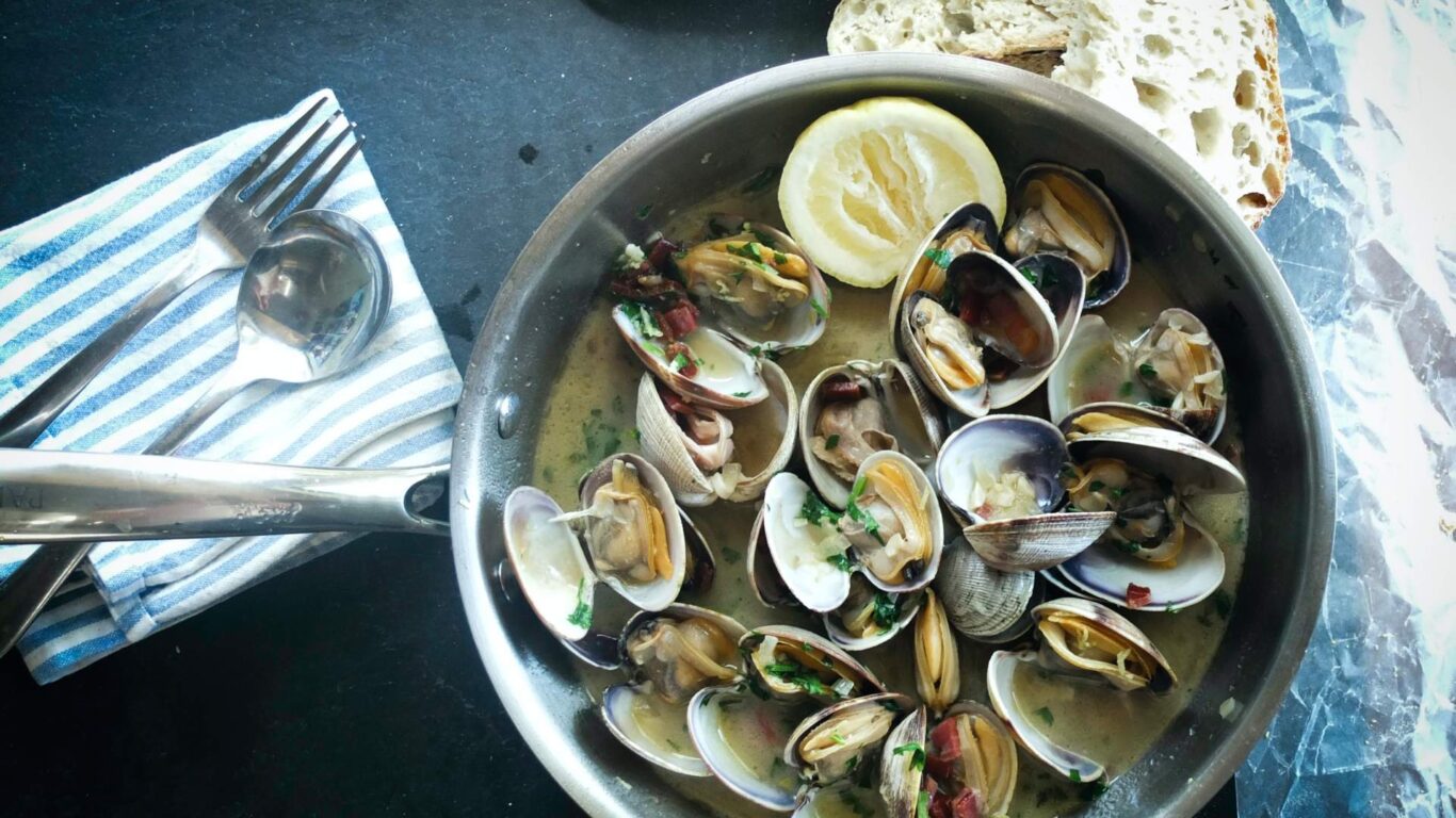 A pan of cooked clams in a broth garnished with herbs and a lemon slice, accompanied by sliced bread, a striped napkin, a spoon, and a fork.