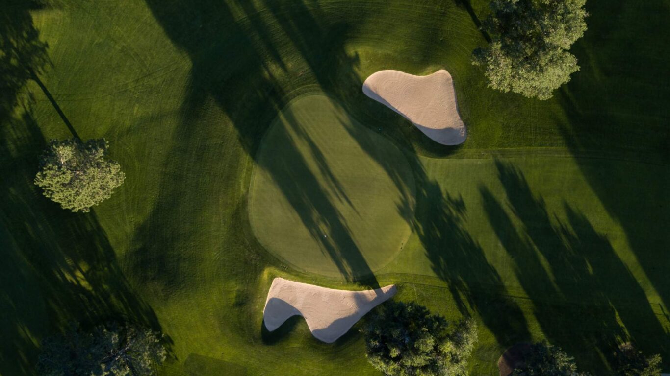 Aerial view of a golf course showing a putting green with two sand bunkers and surrounding trees casting long shadows.