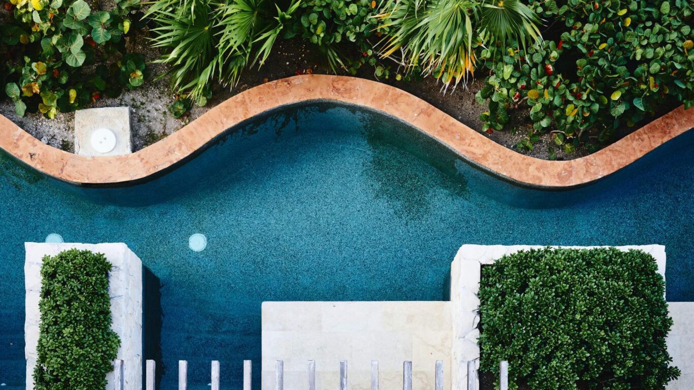 Aerial view of a curving pool with a stone border, surrounded by lush green plants. Steps and a wooden pergola lead to the pool area.