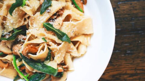 A plate of pasta with mushrooms, fresh greens, and grated cheese, served on a white dish, placed on a dark wooden table.