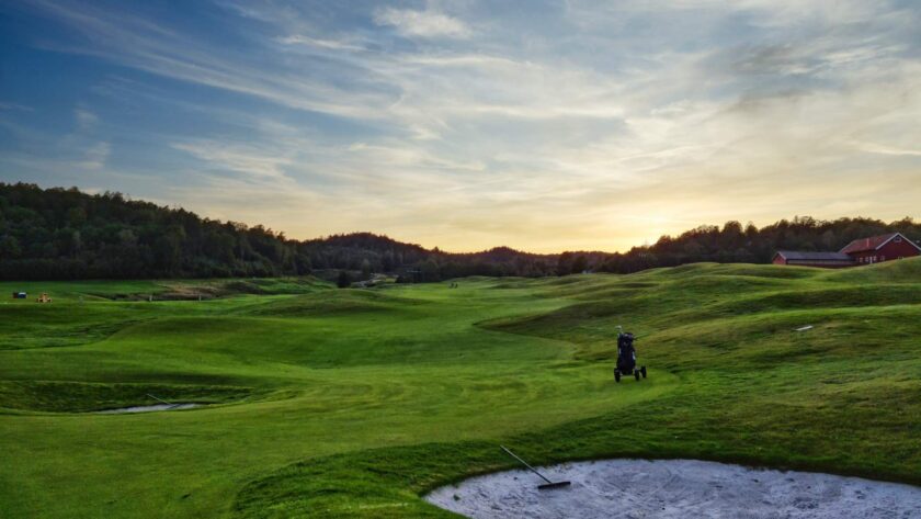 A golf course at sunset with a sand trap in the foreground, a distant golf cart on the fairway, and a mix of trees and buildings in the background under a partly cloudy sky.