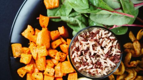 A black plate with cooked brown and white rice in a glass bowl, surrounded by roasted sweet potatoes, spinach leaves, and fried plantains.