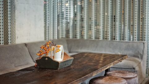 A wooden table with a long gray sofa and two stools. A small wooden tray holding a vase with orange berries is placed on the table. The background features metal paneling.