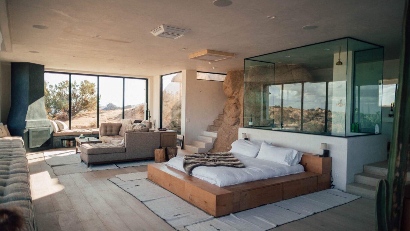 Spacious modern bedroom with a large bed, corner seating area, and floor-to-ceiling windows overlooking a desert landscape. Glass shower enclosure separates bed and seating area.