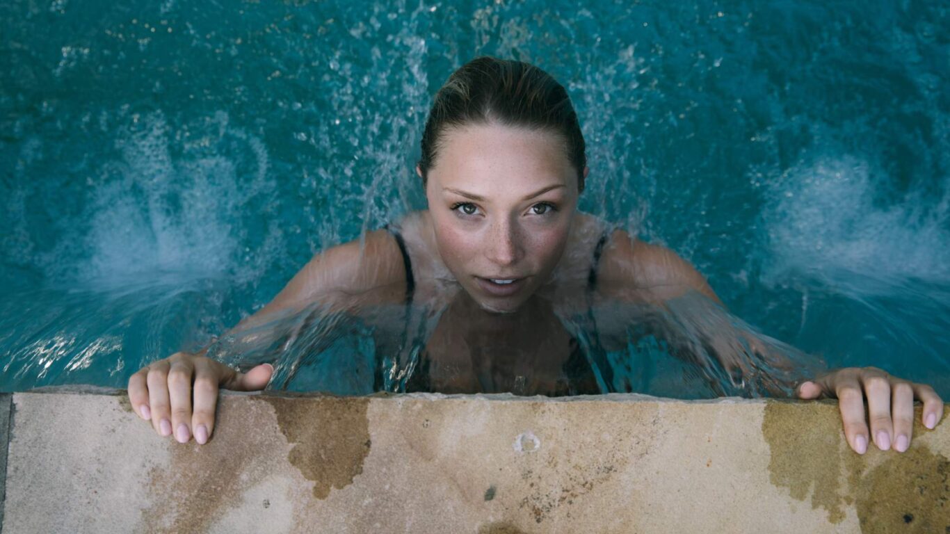 Person in a swimming pool, holding onto the edge, with water cascading over their shoulders.